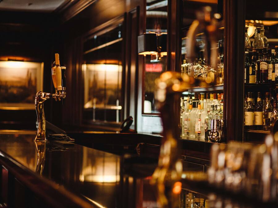 A bar with a polished wooden counter, a tap, and shelves stocked with bottles.