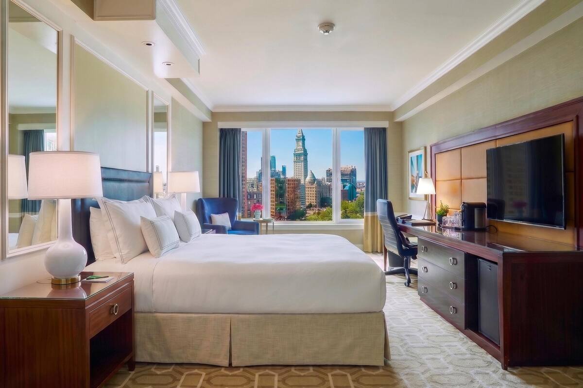 A hotel room with a bed, a desk, a television, and a view of a cityscape.