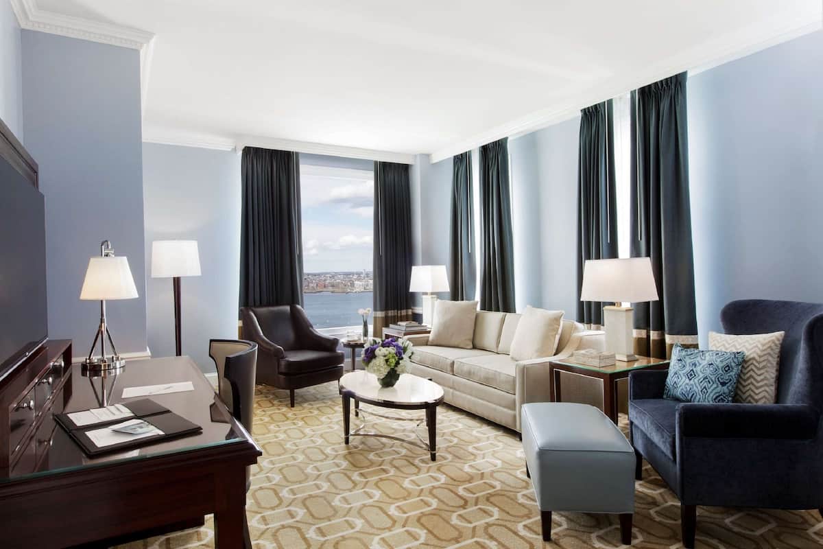 A modern hotel room with a large window, a sofa, a chair, a desk, and a lamp.