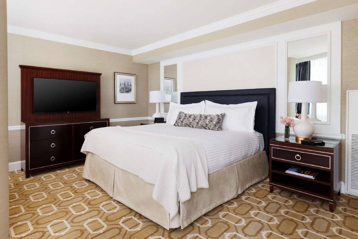 A hotel room with a large bed, a flat-screen TV, a dresser, and a nightstand with a lamp.