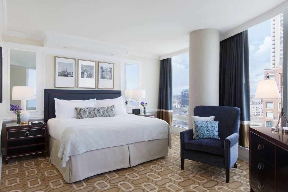 A hotel room with a large bed, a sitting area with a chair, a nightstand, a lamp, and a view of the city through large windows.