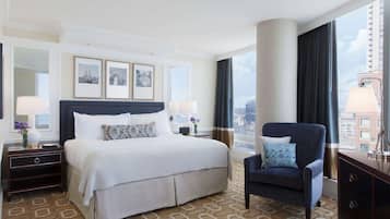 Ein Hotelzimmer mit einem großen Bett, einer Sitzecke mit einem Sessel, einem Nachttisch, einer Lampe und Blick auf die Stadt durch große Fenster.