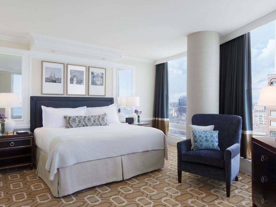 Ein Hotelzimmer mit einem großen Bett, einer Sitzecke mit einem Sessel, einem Nachttisch, einer Lampe und Blick auf die Stadt durch große Fenster.