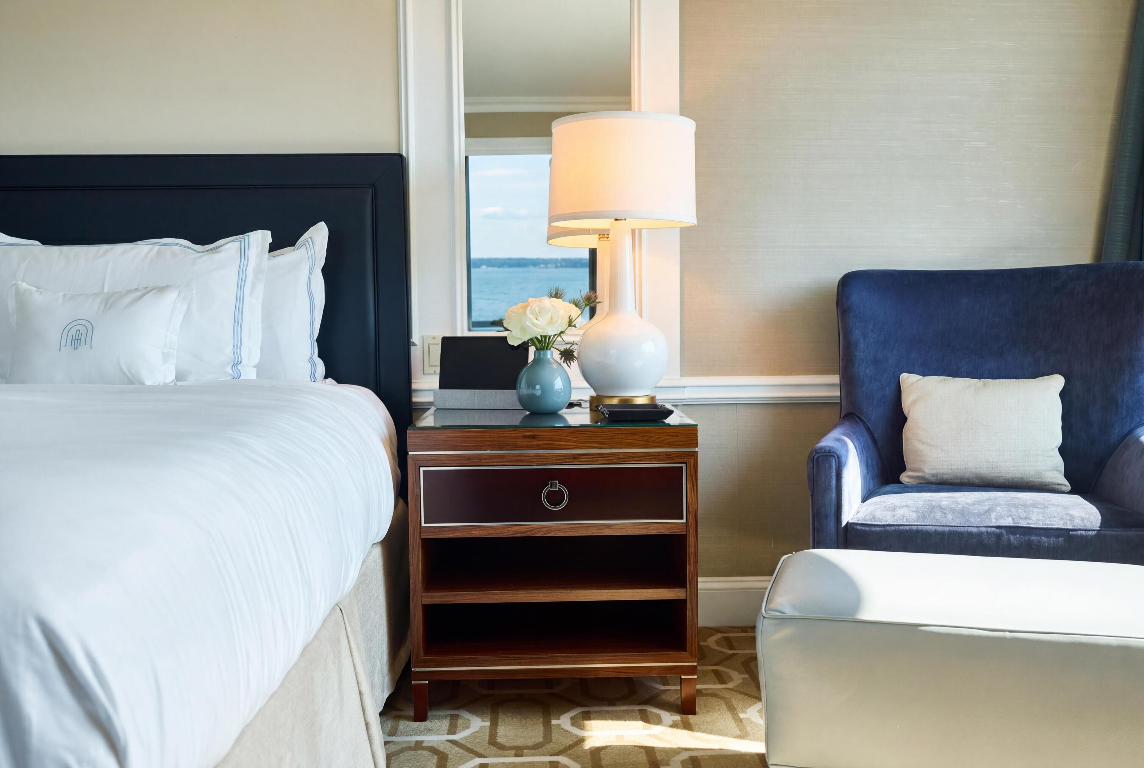 Quarto de hotel com uma cama, mesa de cabeceira com luminária, uma cadeira e vista para o mar.