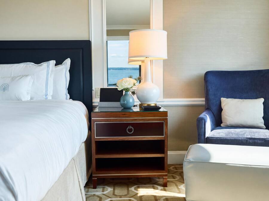 Quarto de hotel com uma cama, mesa de cabeceira com luminária, uma cadeira e vista para o mar.