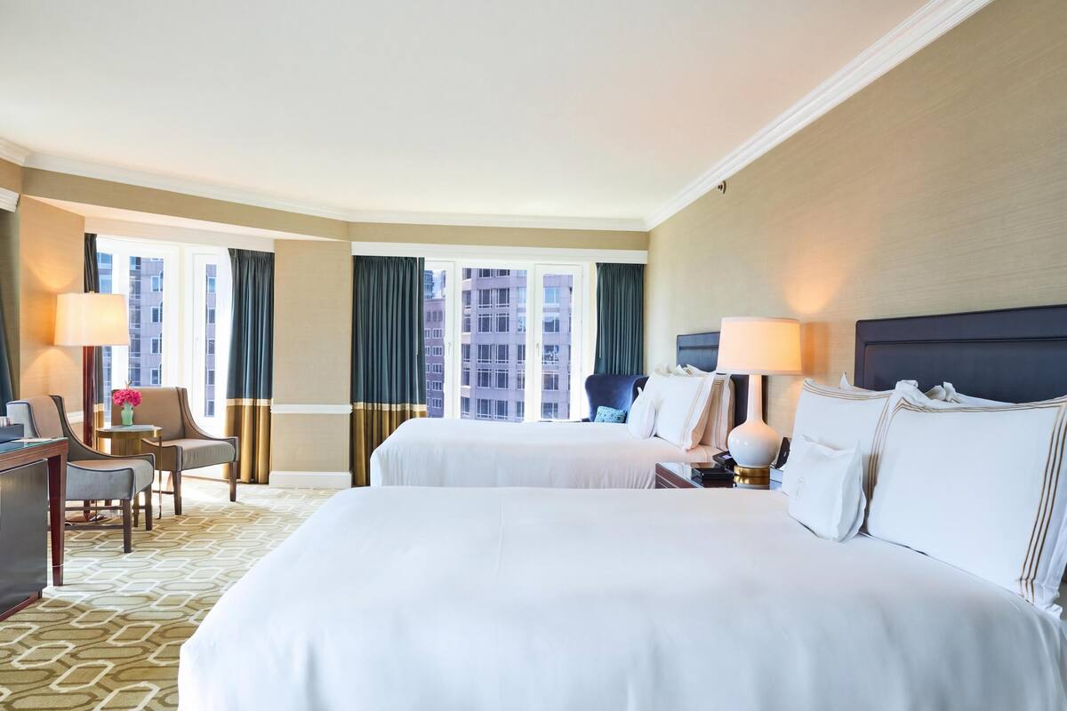 A hotel room with two beds, a desk, a chair, a lamp, and a view of buildings through large windows.