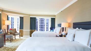 A hotel room with two beds, a desk, a chair, a lamp, and a view of buildings through large windows.