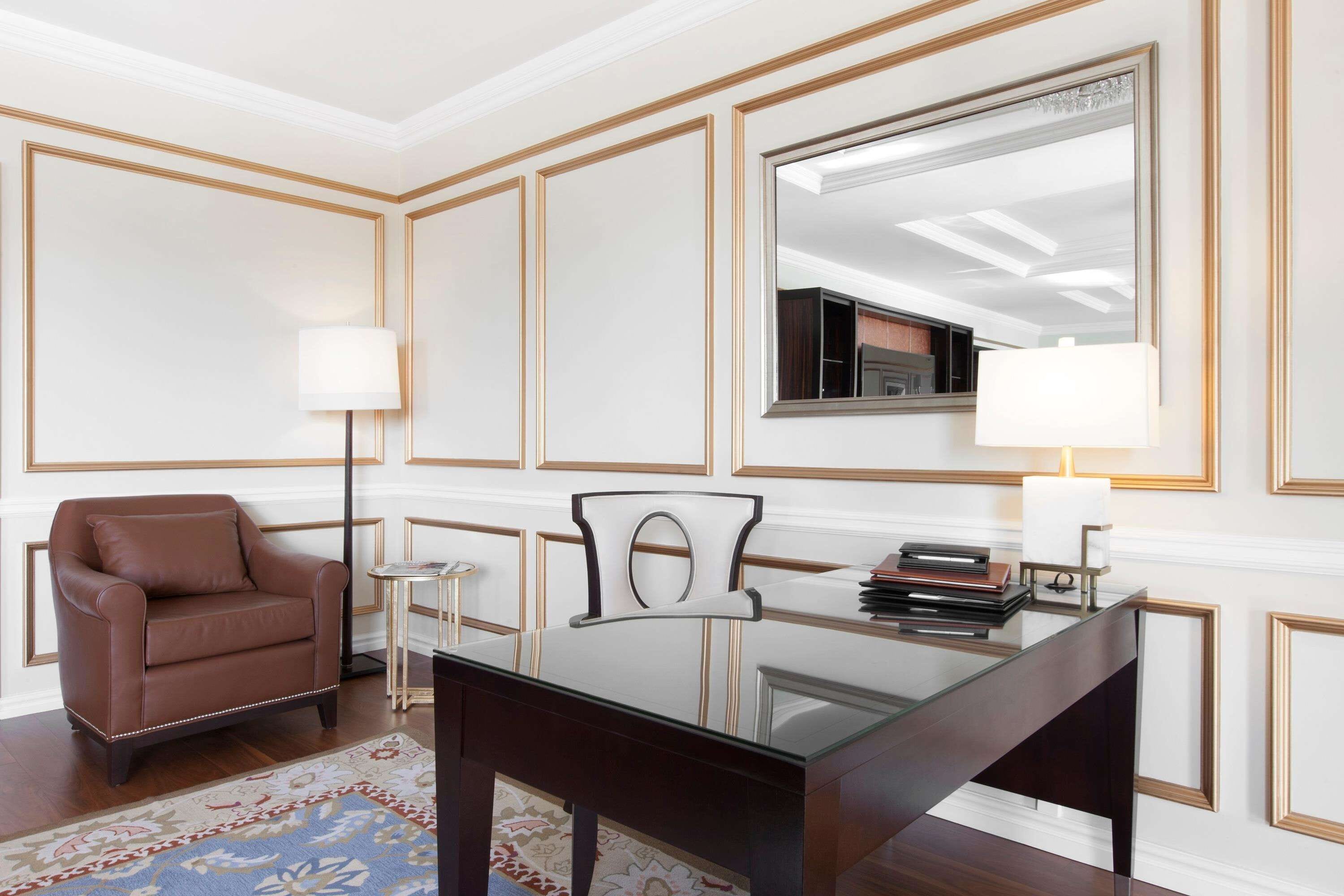 Una habitación de hotel moderna con un escritorio de vidrio, un sillón de cuero, una mesita auxiliar, una lámpara y un espejo en la pared.