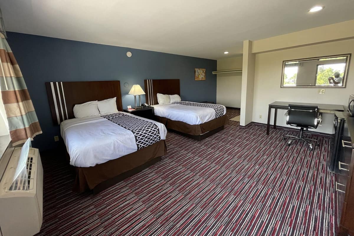 Quarto de hotel com duas camas, uma escrivaninha, uma cadeira, um abajur e uma janela.