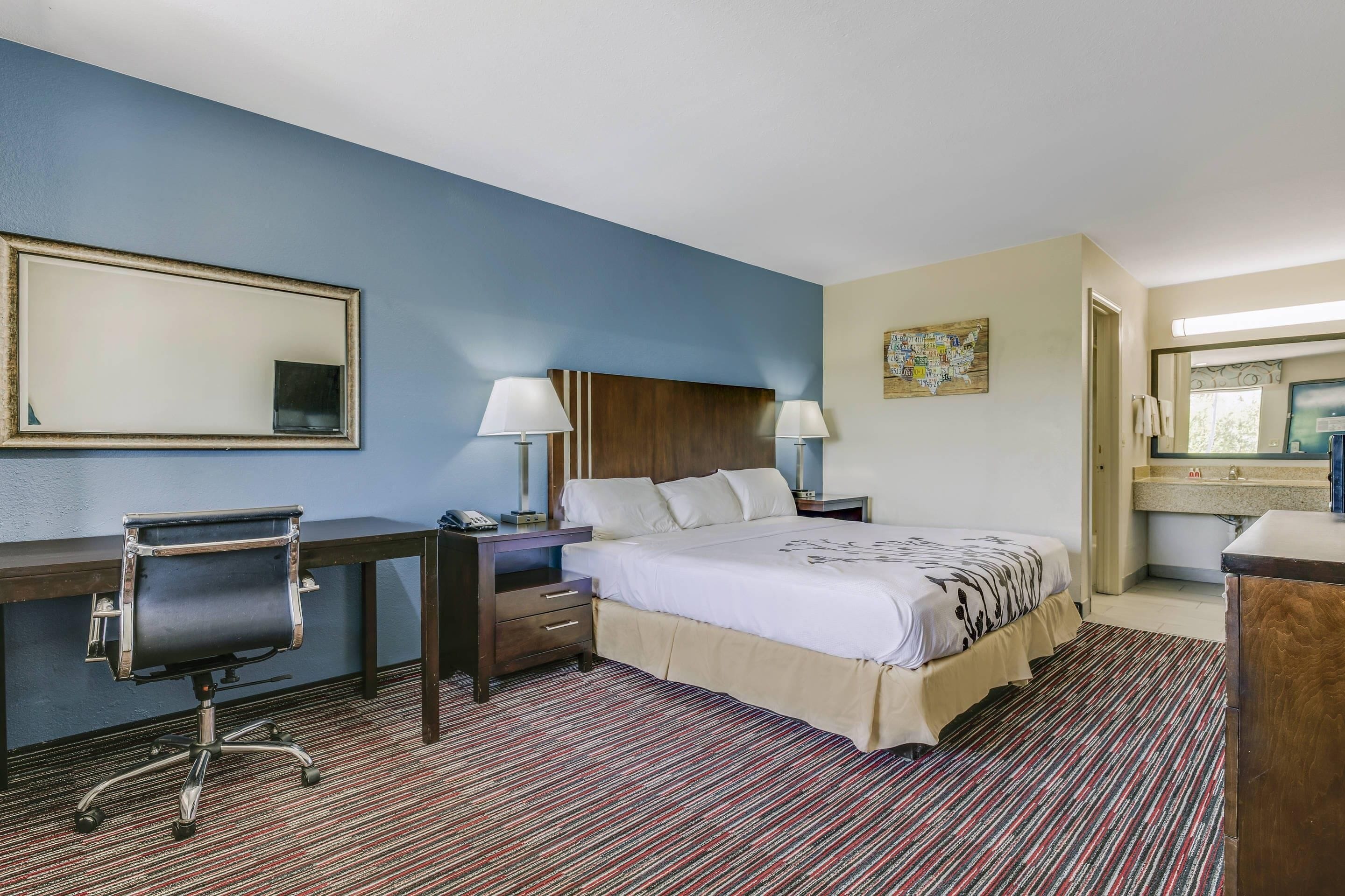 Quarto de hotel com cama, escrivaninha, cadeira e banheiro visível por uma porta aberta.