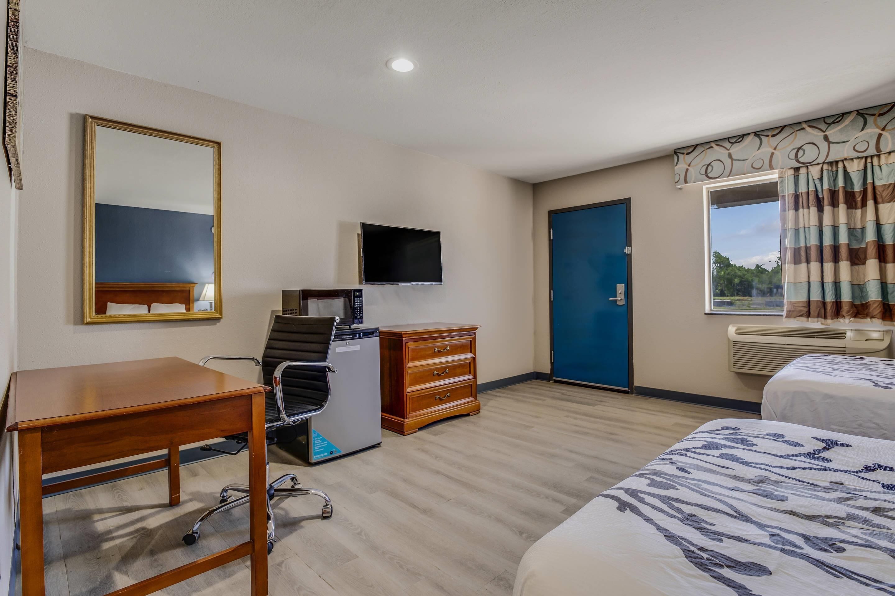 A hotel room with a desk, chair, dresser, bed, TV, and a window with curtains.