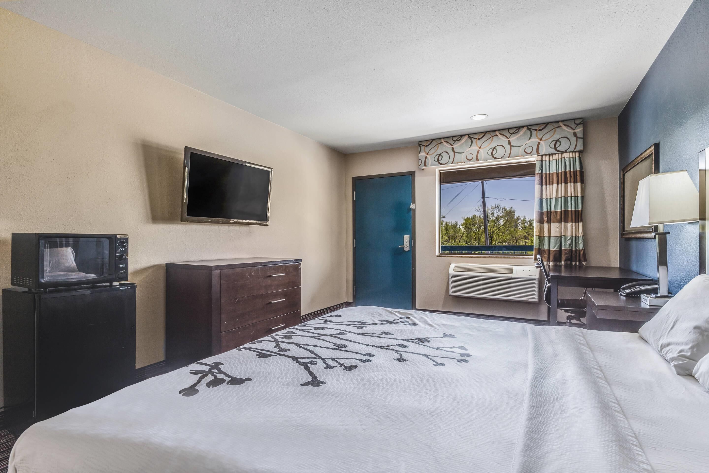 A hotel room with a bed, a TV mounted on the wall, a desk with a lamp, and a window with curtains.