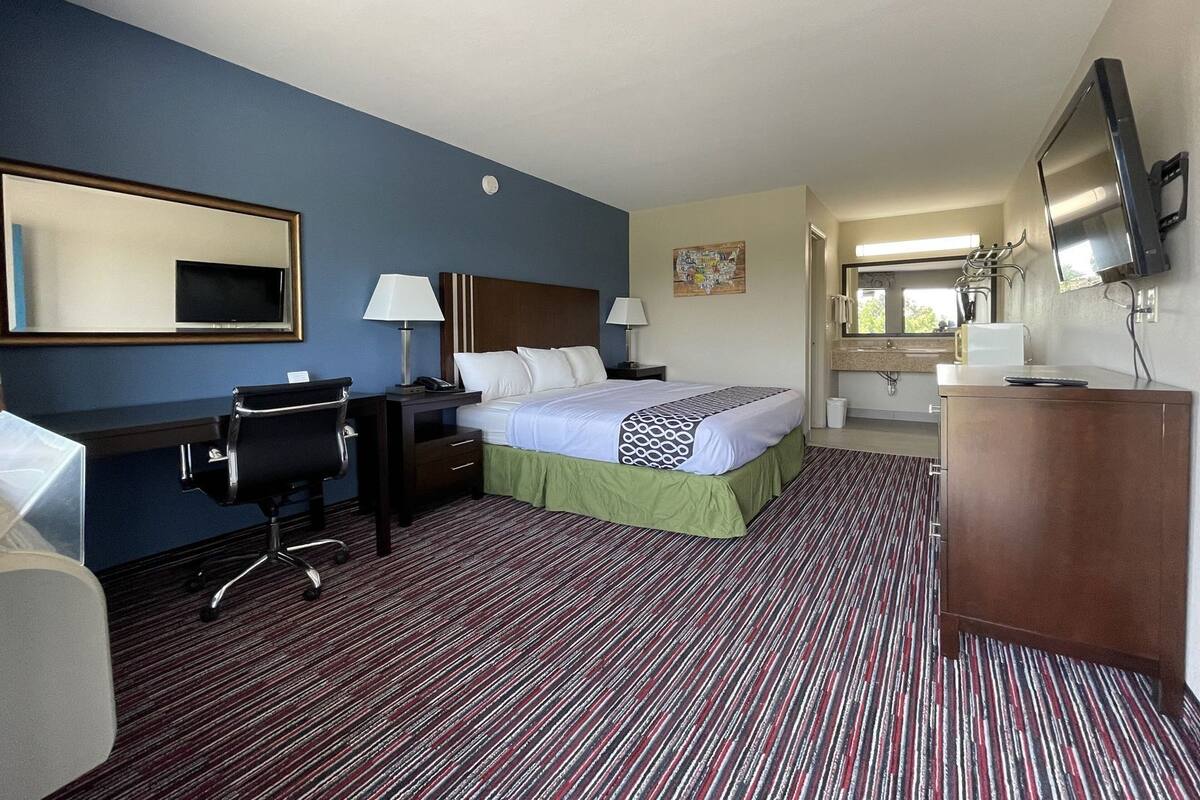 Quarto de hotel com cama, escrivaninha, cadeira, televisão e banheiro com espelho e pia.