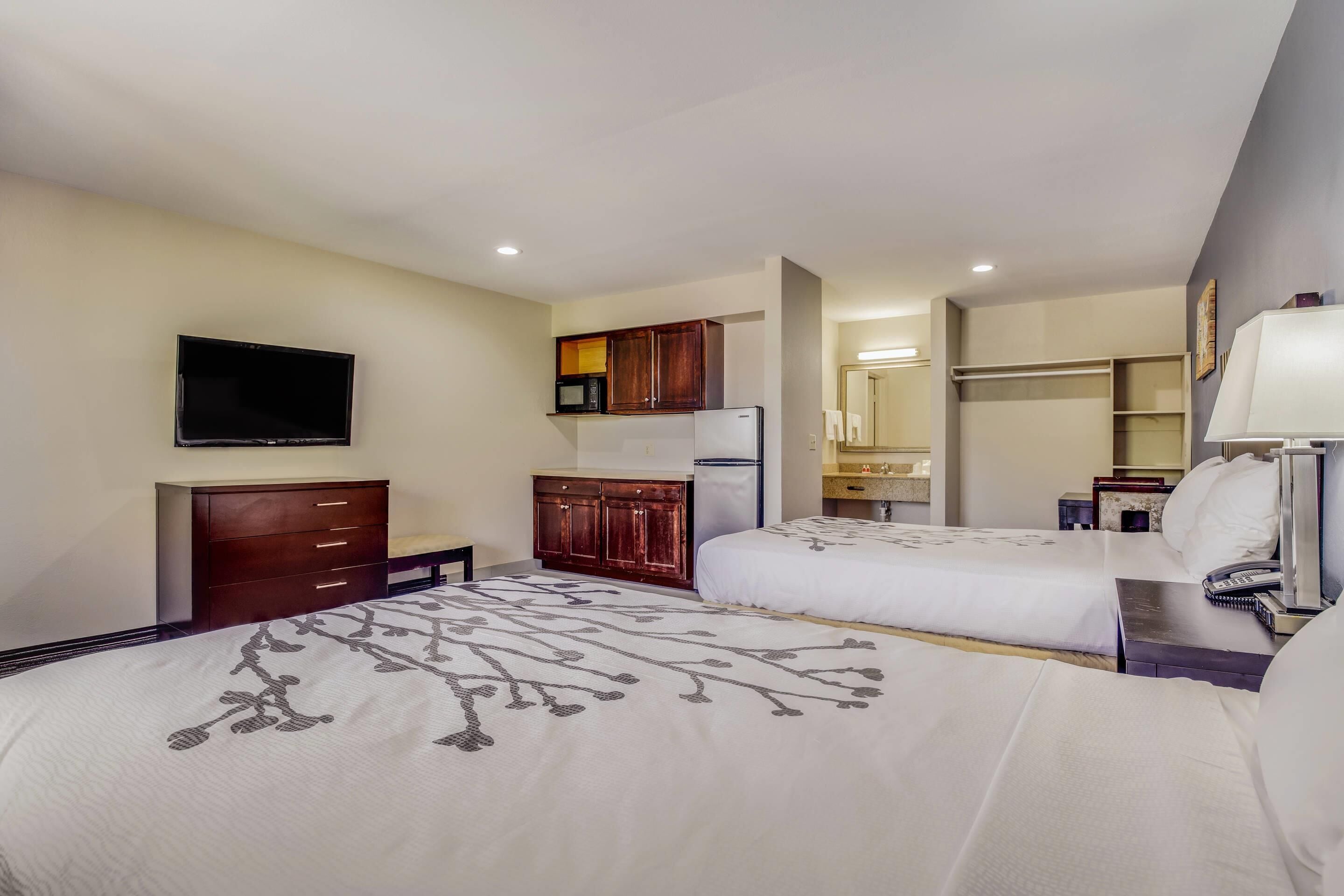 A hotel room with two beds, a TV, a microwave, a refrigerator, a desk with a lamp, and a bathroom visible through an open door.