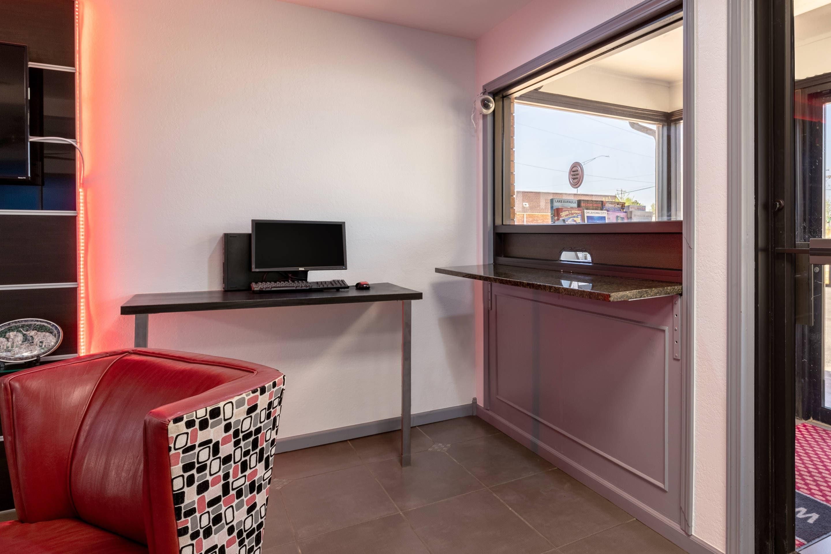 Una habitación con escritorio, computadora, una silla roja y ventana con vista a un edificio.