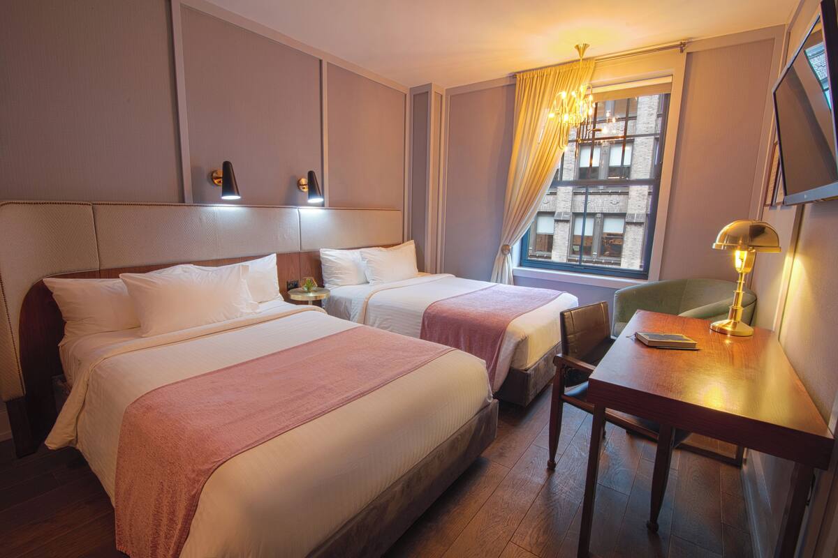A hotel room with two beds, a desk, a chair, a lamp, a window with curtains, and a view of a building.