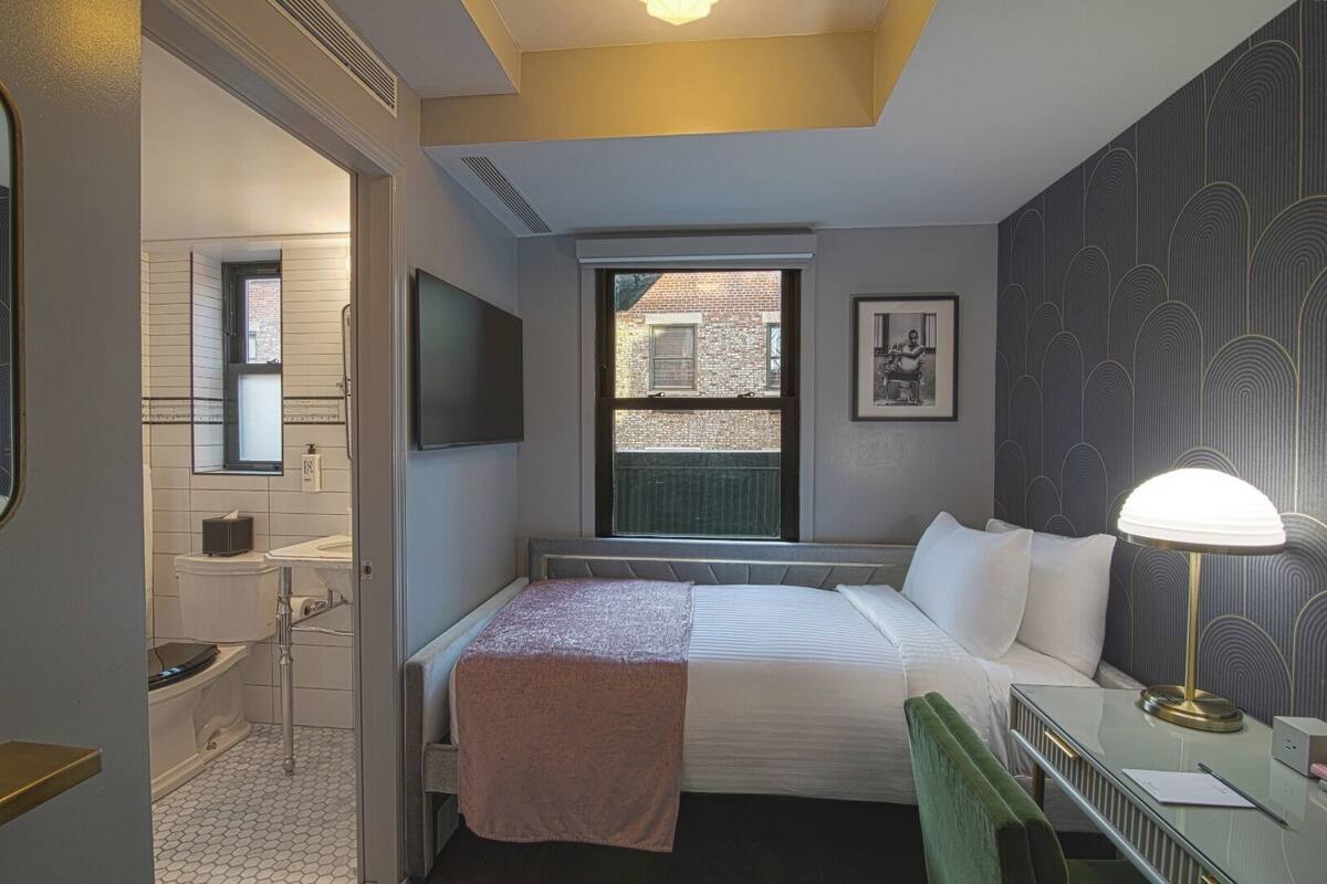 A hotel room with a bed, bedside table, lamp, and a window with a view of the outside.