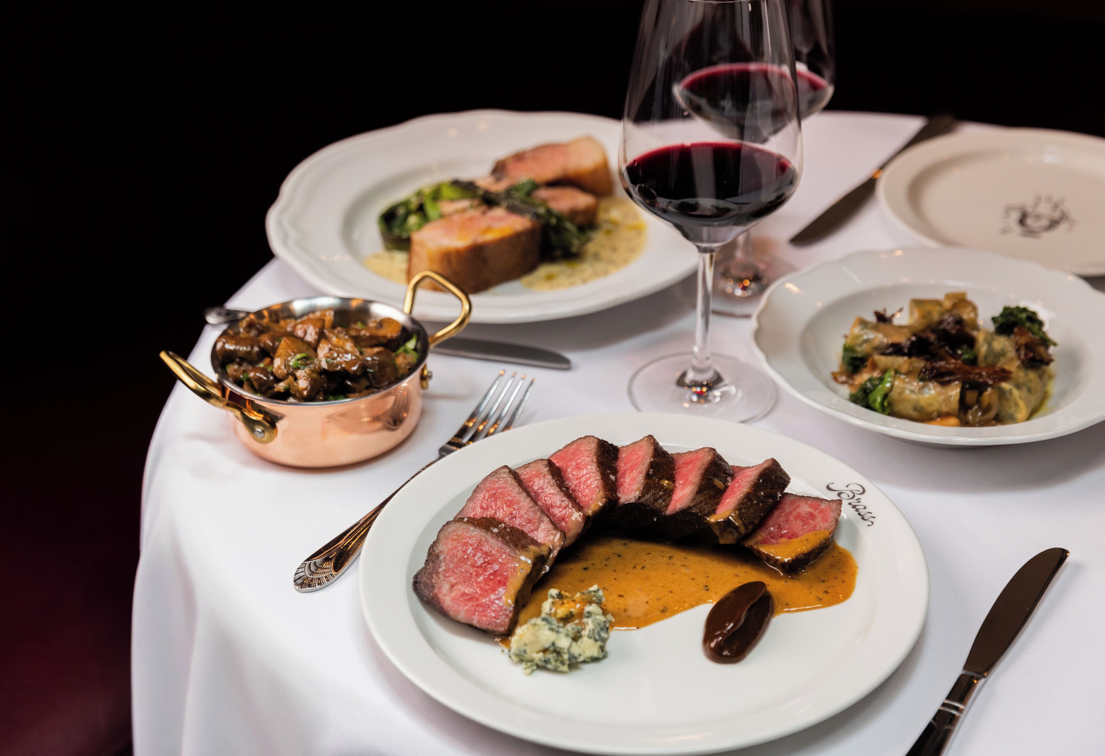 A table set with multiple plates of food, including steak and vegetables, and a glass of red wine.