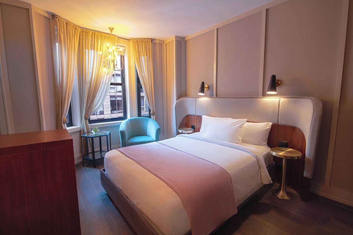 A hotel room with a large bed, a desk, a chair, a small table, and a window with curtains.