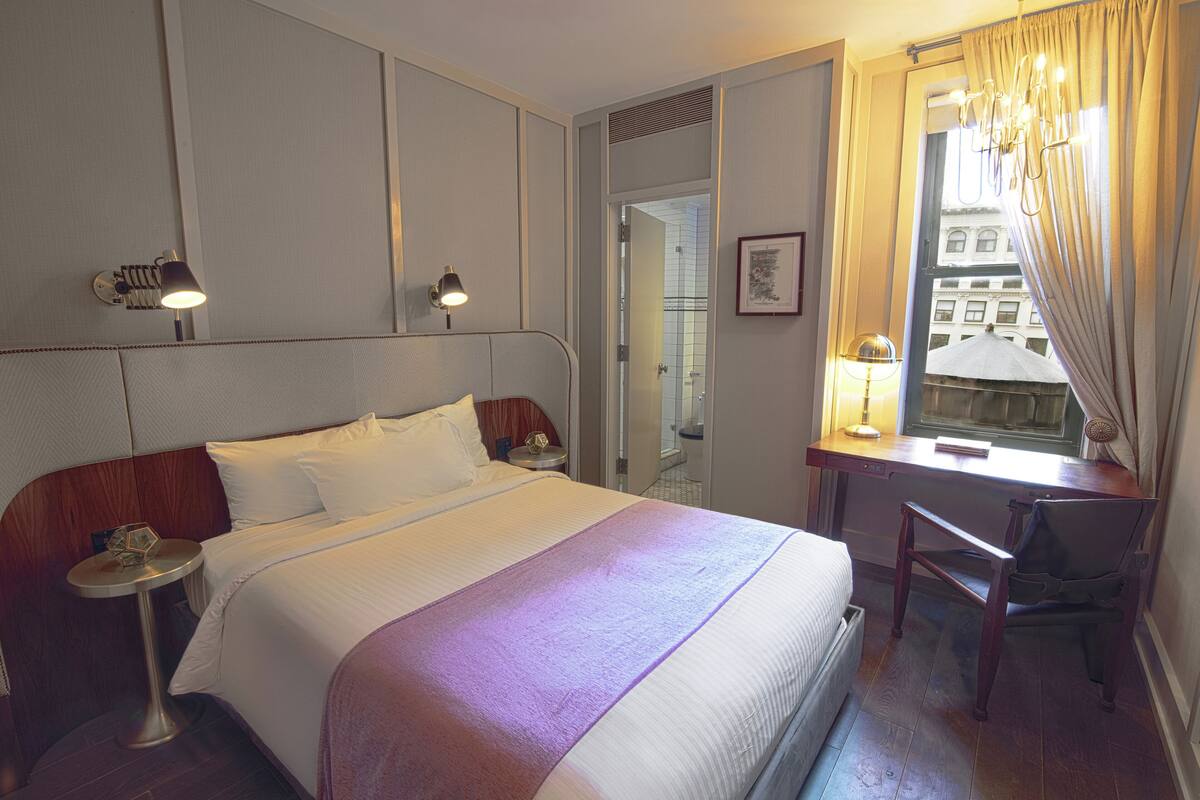 A hotel room with a bed, bedside tables, a desk with a chair, a window with curtains, and a view of buildings outside.
