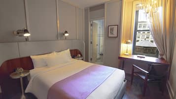 A hotel room with a bed, bedside tables, a desk with a chair, a window with curtains, and a view of buildings outside.