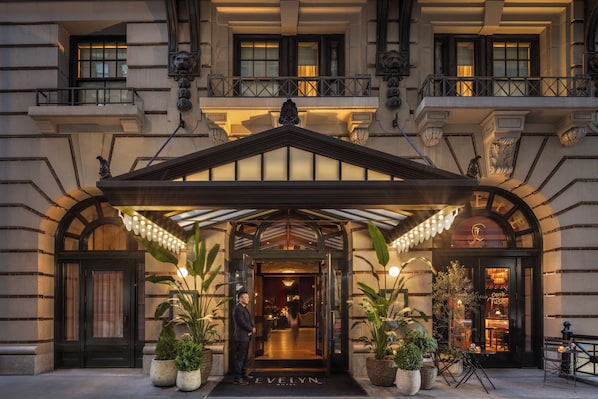 Entrada de hotel con marquesina, plantas en macetas y letrero de restaurante a la vista.