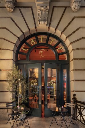 A café with a classic arched entrance, outdoor seating with tables and chairs, and a visible interior through the glass doors. - The Evelyn Hotel (New York)
