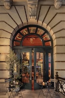 A café with a classic arched entrance, outdoor seating with tables and chairs, and a visible interior through the glass doors.