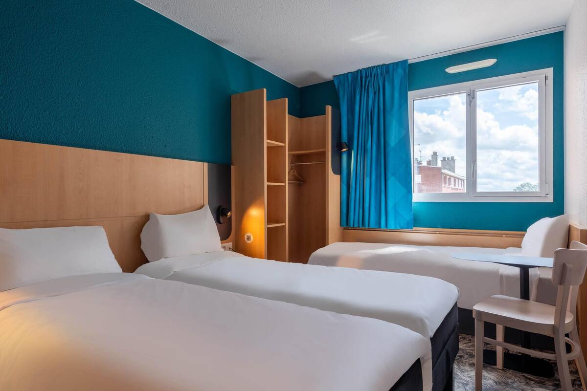 Habitación de hotel con dos camas, un armario de madera, un escritorio, una silla y una ventana con vistas a edificios.