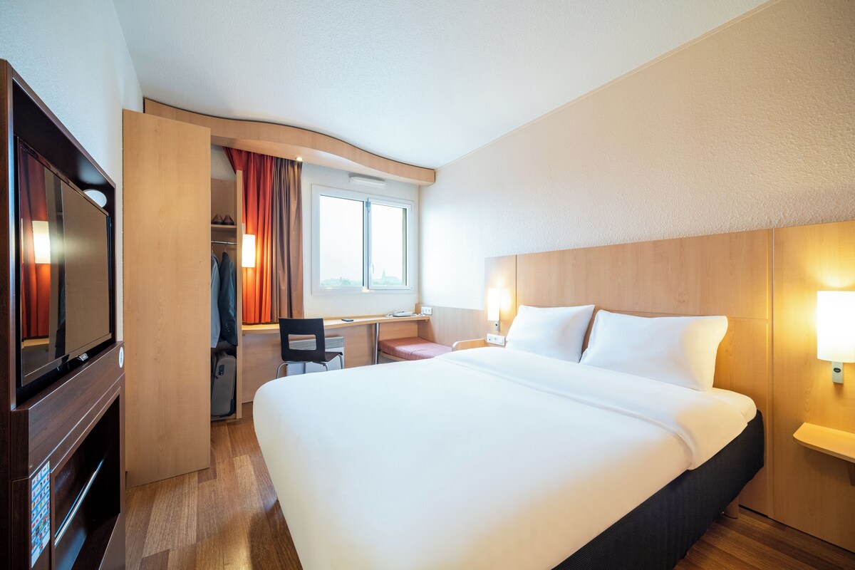 Habitación de hotel con una cama grande, un escritorio con silla, televisión y ventana con cortinas.