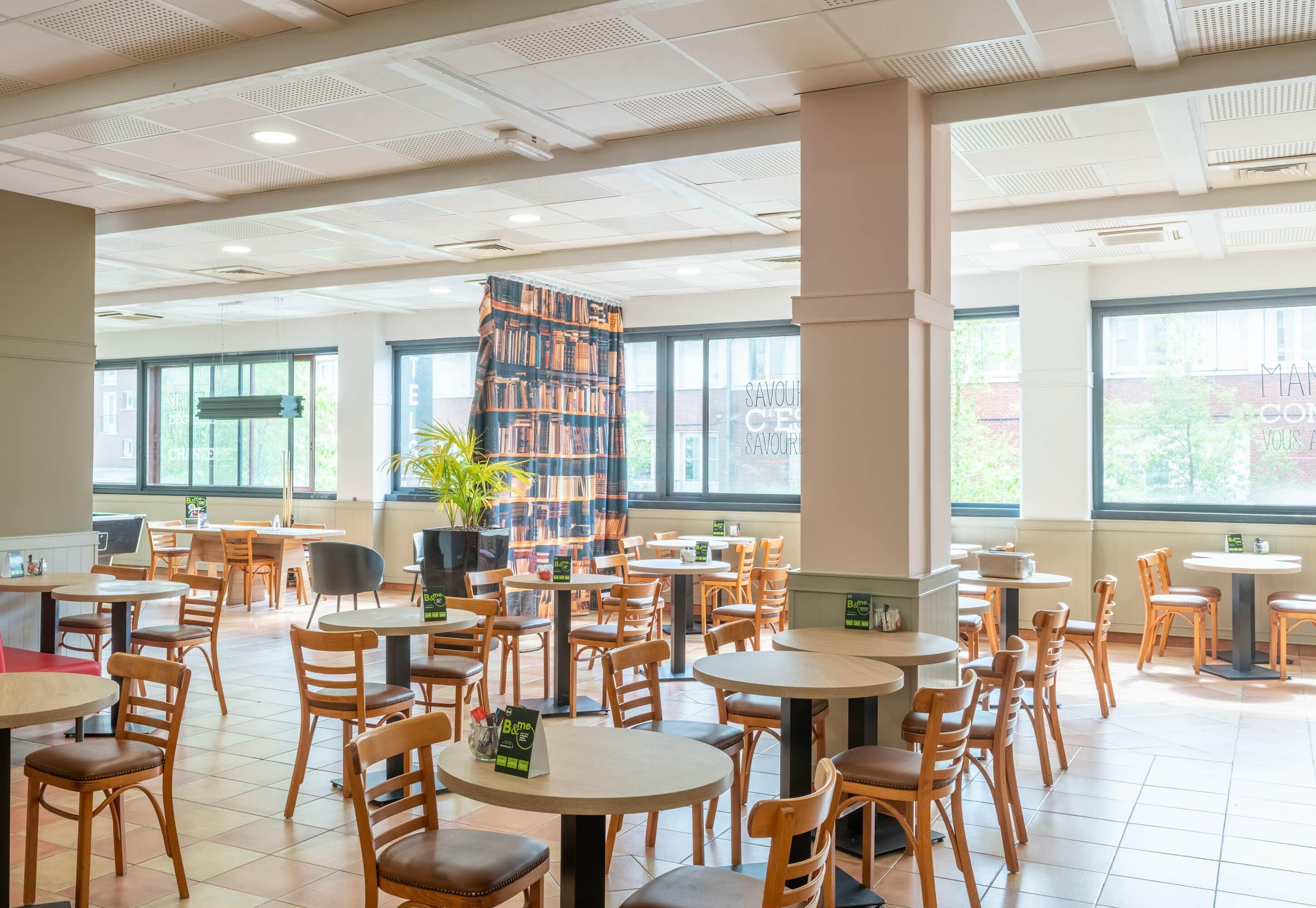 A spacious indoor dining area with multiple tables and chairs, large windows, and a central pillar with a book-covered design.