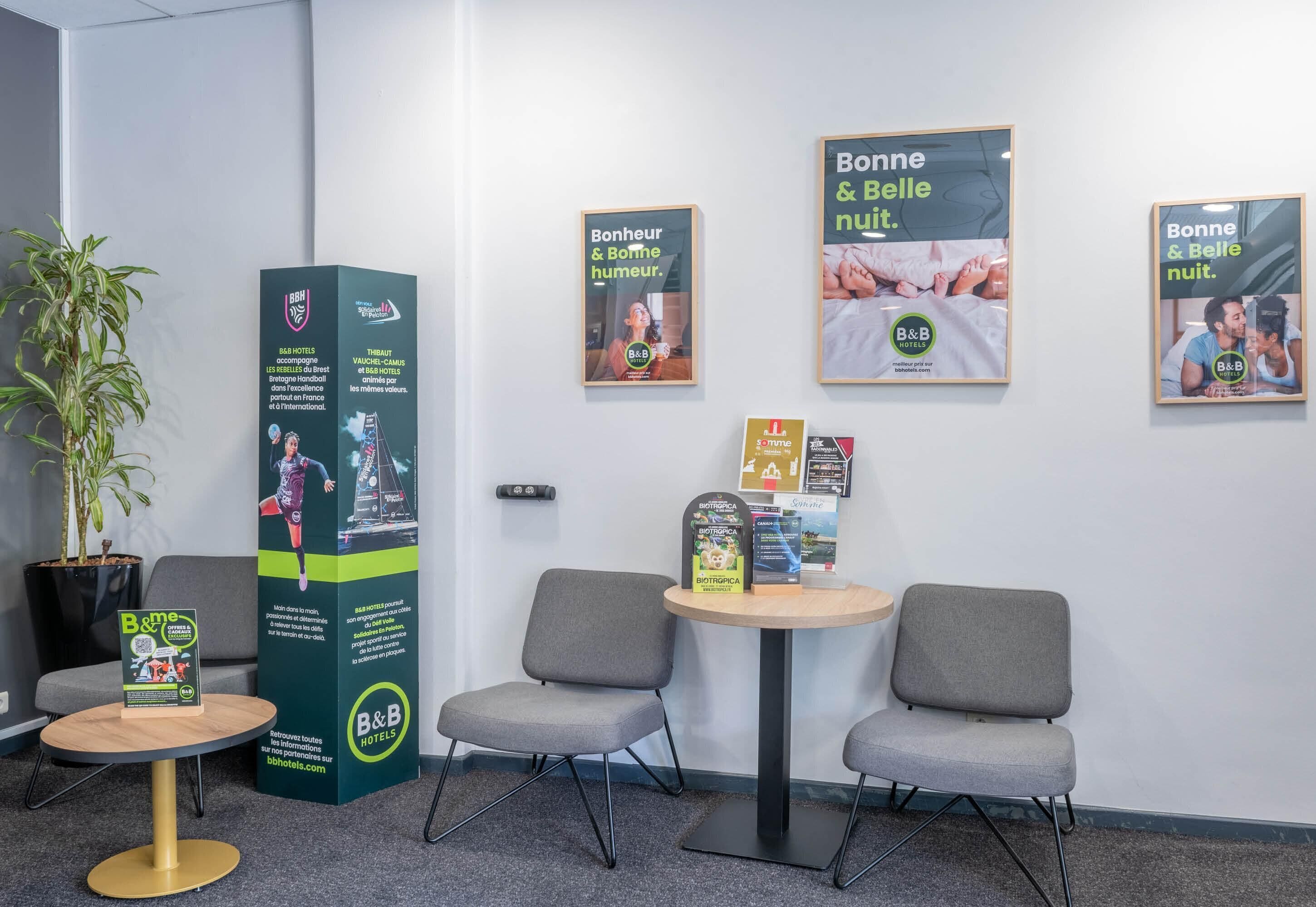 Een receptiegedeelte met een potplant, een hoge informatiezuil, twee grijze stoelen, een kleine ronde tafel en ingelijste posters aan de muur.