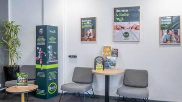 Een receptiegedeelte met een potplant, een hoge informatiezuil, twee grijze stoelen, een kleine ronde tafel en ingelijste posters aan de muur.