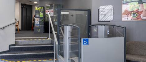 Entrada de hotel con escalera, ascensor accesible para sillas de ruedas y mostrador de recepción.