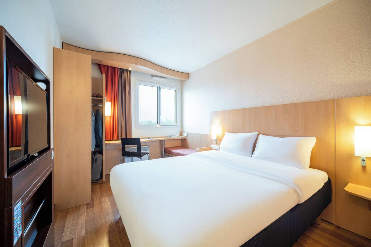 Habitación de hotel con una cama grande, un escritorio con silla, televisión y ventana con cortinas.