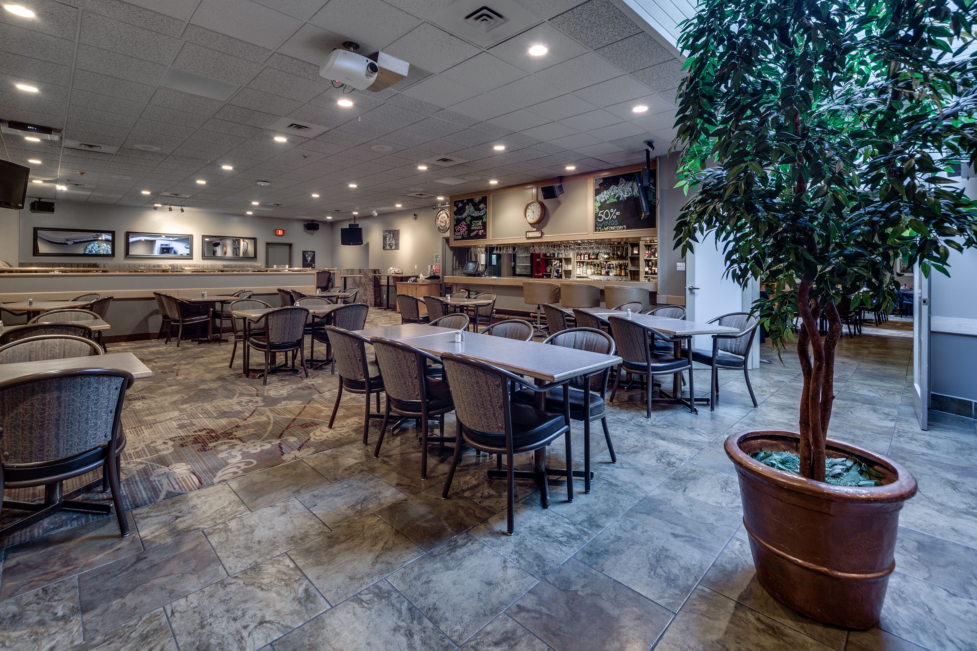 A restaurant with multiple tables and chairs, a bar area, and a large potted tree.