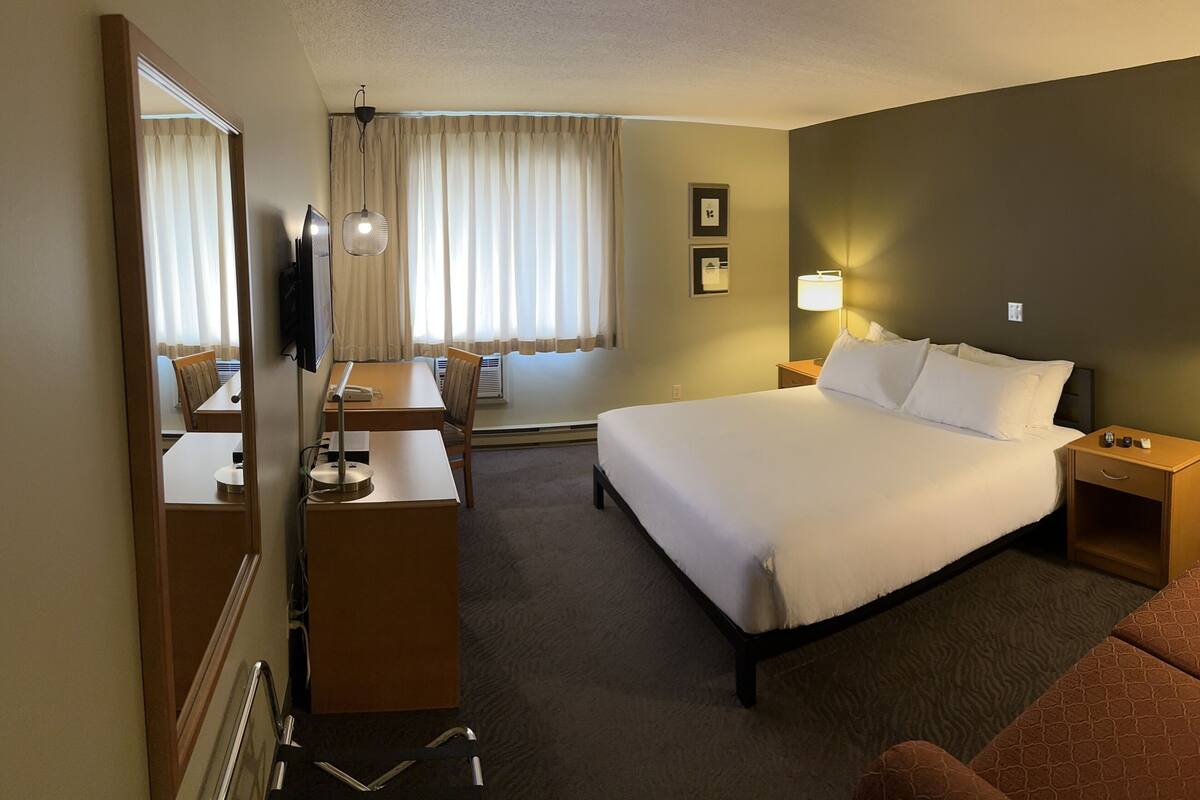 Quarto de hotel com cama, escrivaninha, cadeira, televisão, abajur e janela com cortinas.