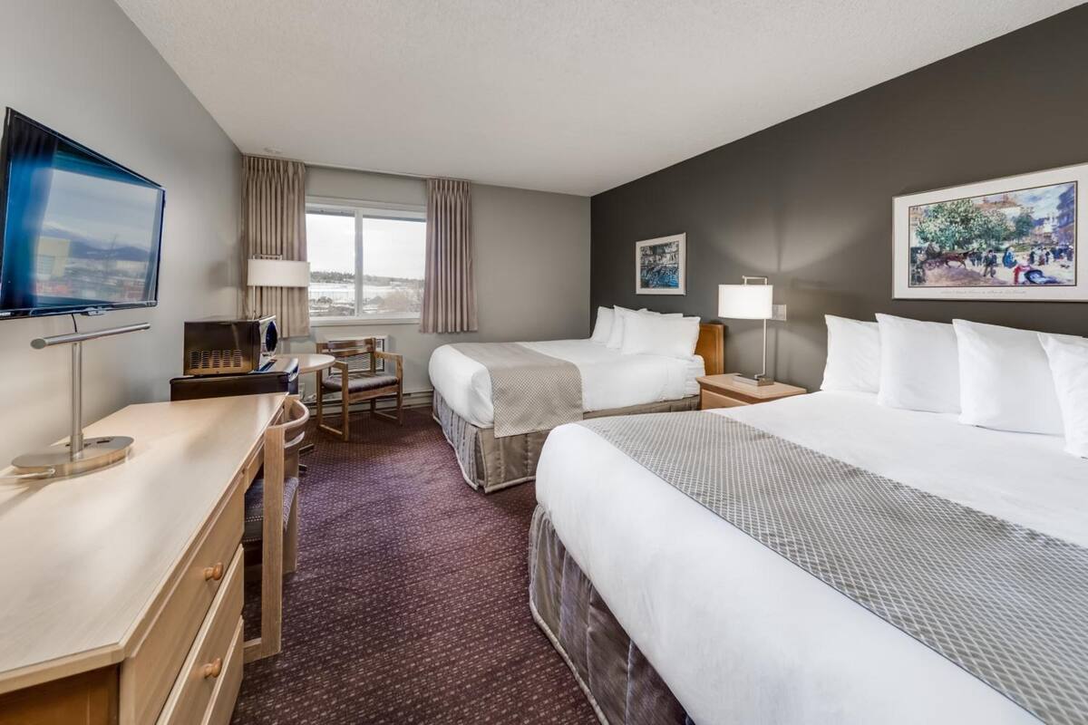 Quarto de hotel com duas camas, uma escrivaninha, televisão e janela com cortinas.