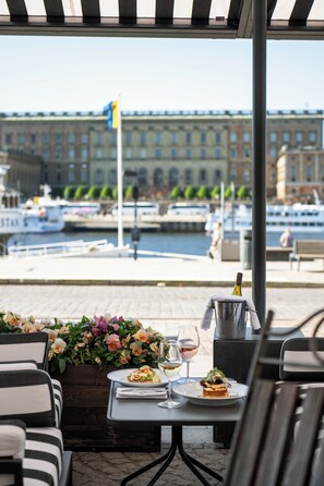 An outdoor dining area with tables and chairs, a view of a marina with boats, and a building with a flag in the background. - Lydmar Hotel (Stockholm)