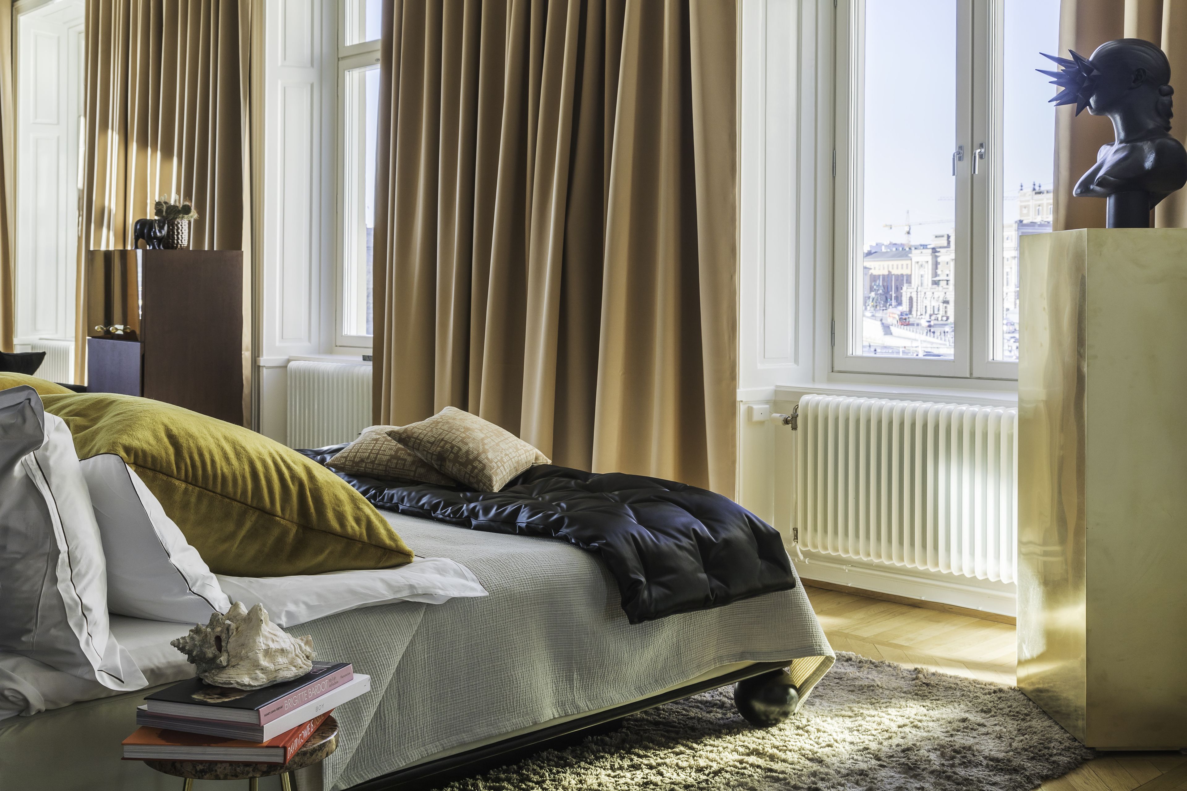 Une chambre à coucher avec un lit, une fenêtre avec des rideaux, un radiateur et un buste posé sur une commode.