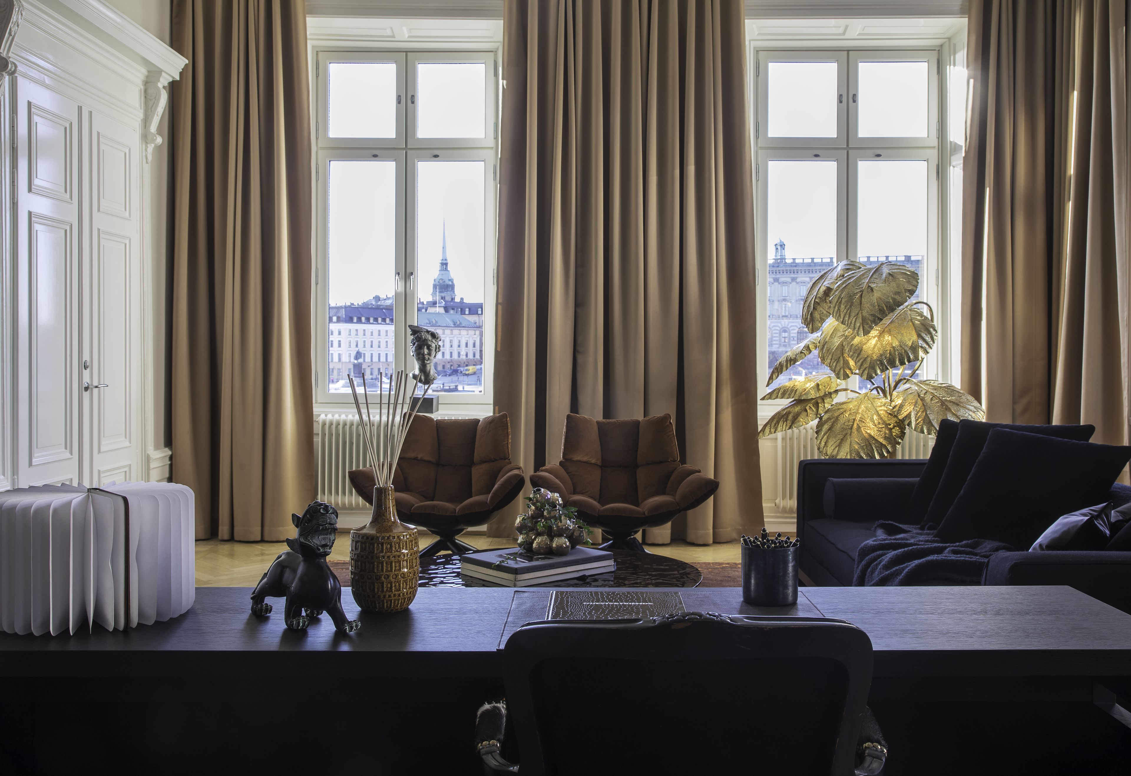 A room with a view of a cityscape, featuring a large window, a sofa, a table with a vase and a book, and a radiator.