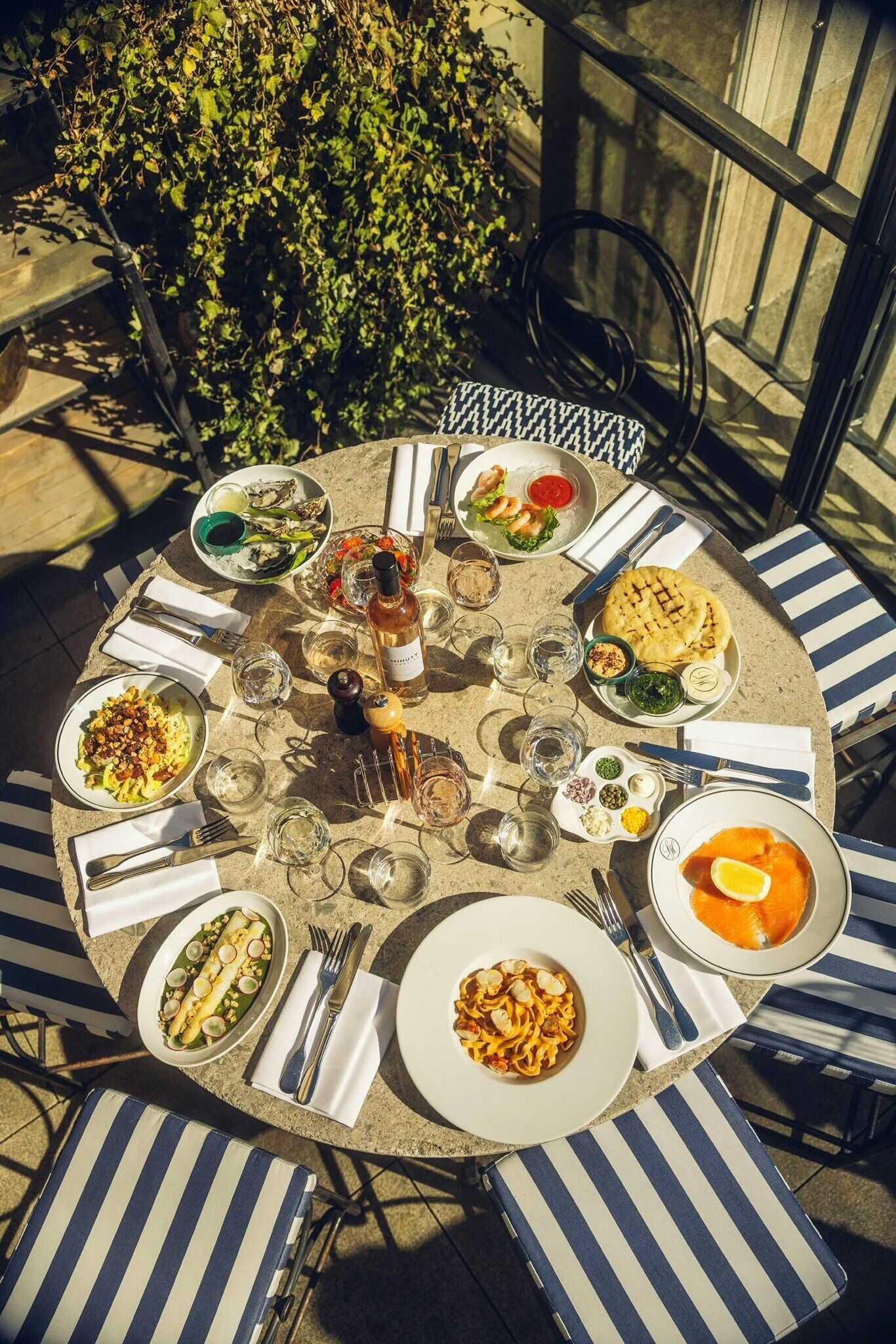 Un tavolo da pranzo all'aperto a forma di cerchio con sedie a righe, circondato da piatti e bicchieri, con sullo sfondo piante e un edificio.