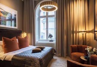 A hotel room with a large bed, a sitting area with a chair, a table with a vase of flowers, and a window with curtains.