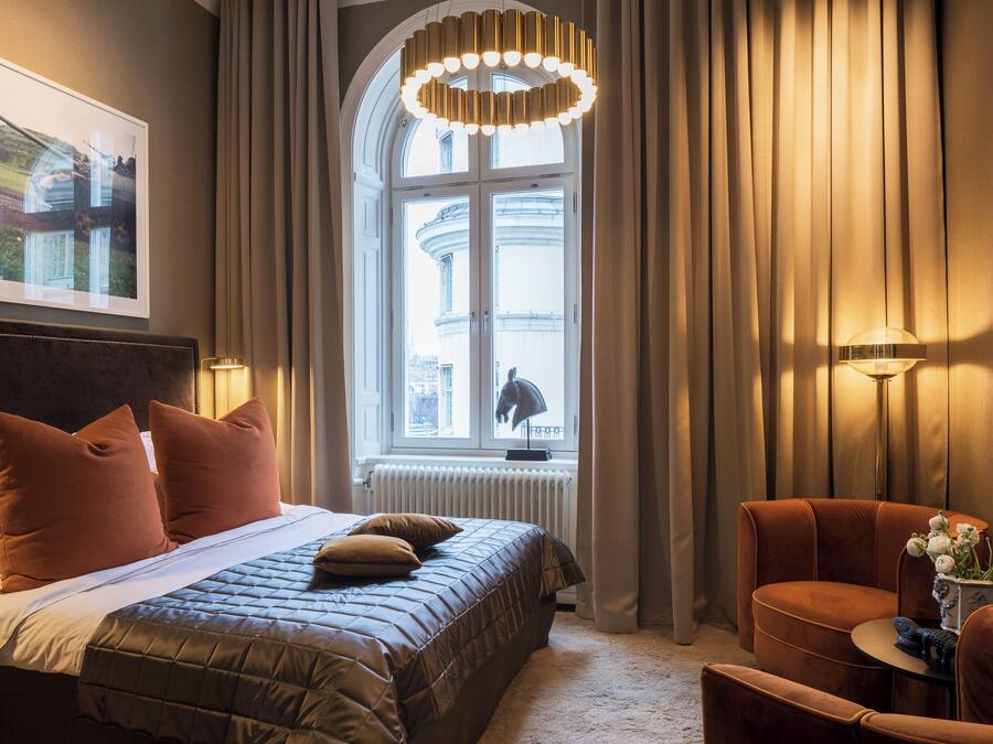 Ett hotellrum med en stor säng, en sittgrupp med en fåtölj, ett bord med en vas med blommor och ett fönster med gardiner.