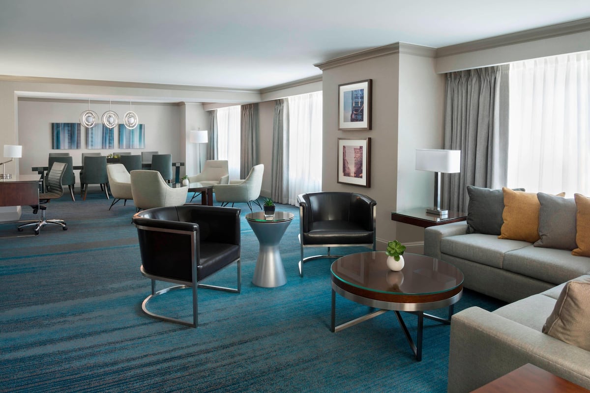 A hotel room with a blue carpet, a sofa, a coffee table, a dining area with chairs, and a TV.