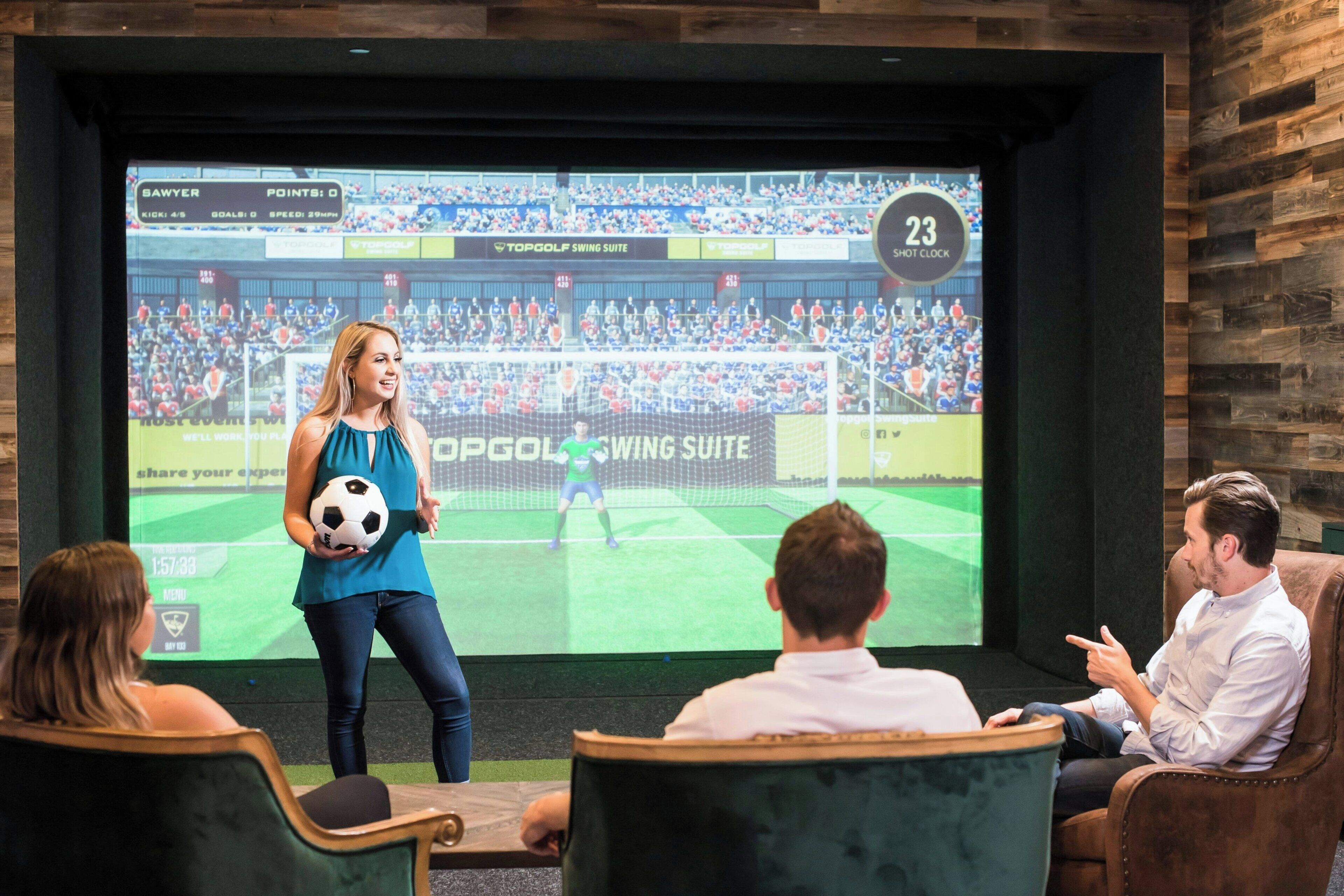 A room with a large screen displaying a soccer game, a woman holding a soccer ball, and people seated watching.