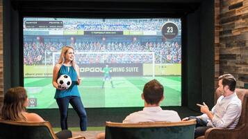 A room with a large screen displaying a soccer game, a woman holding a soccer ball, and people seated watching.