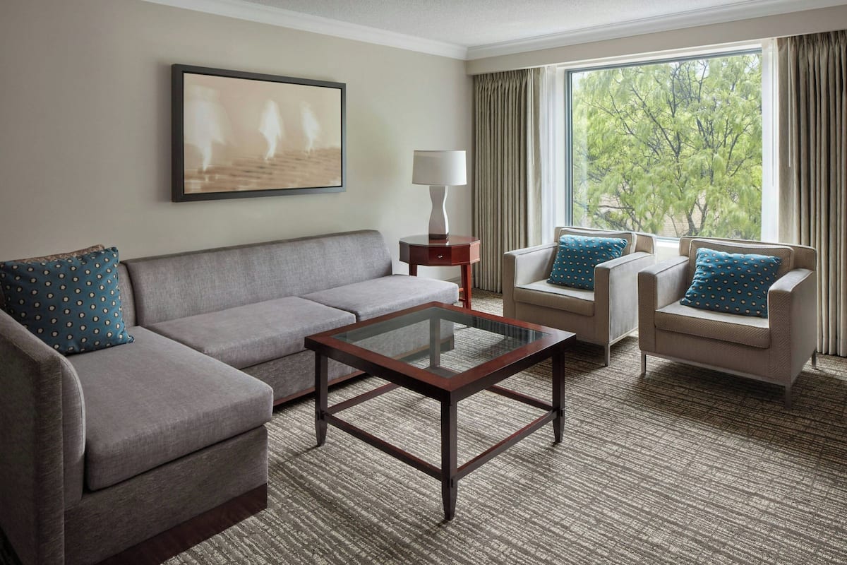 A hotel room with a grey sofa, a glass-top coffee table, a lamp, and two armchairs with patterned cushions. A painting is on the wall, and there is a window with curtains.