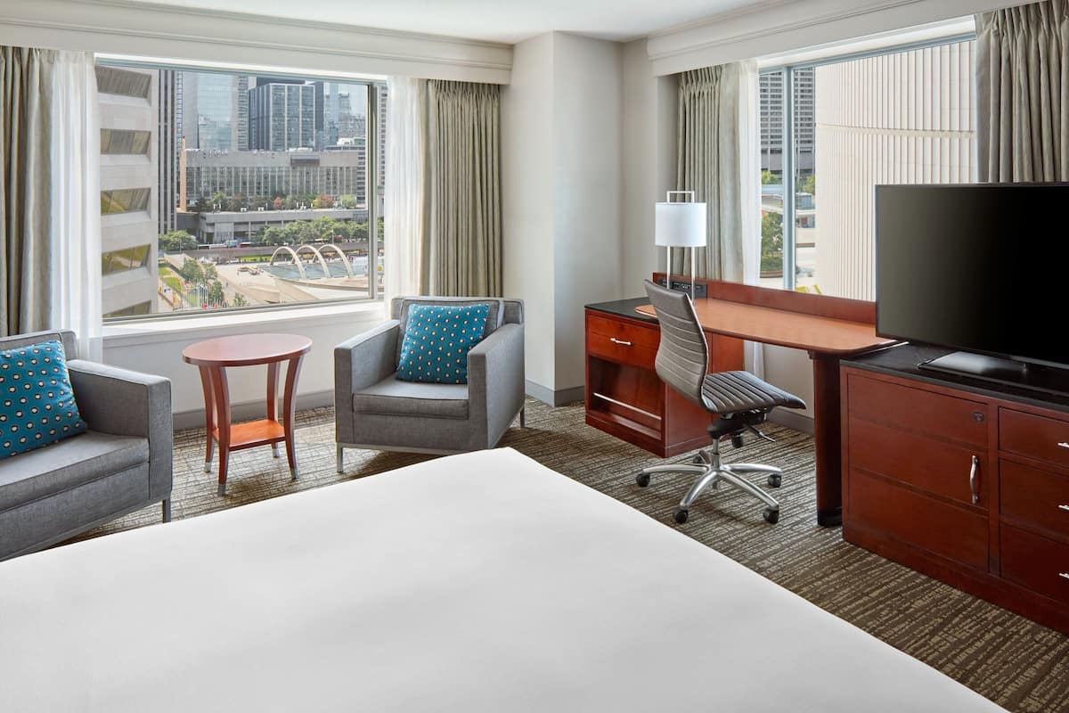 A hotel room with a large bed, a desk with a TV, two armchairs, and a view of the cityscape.