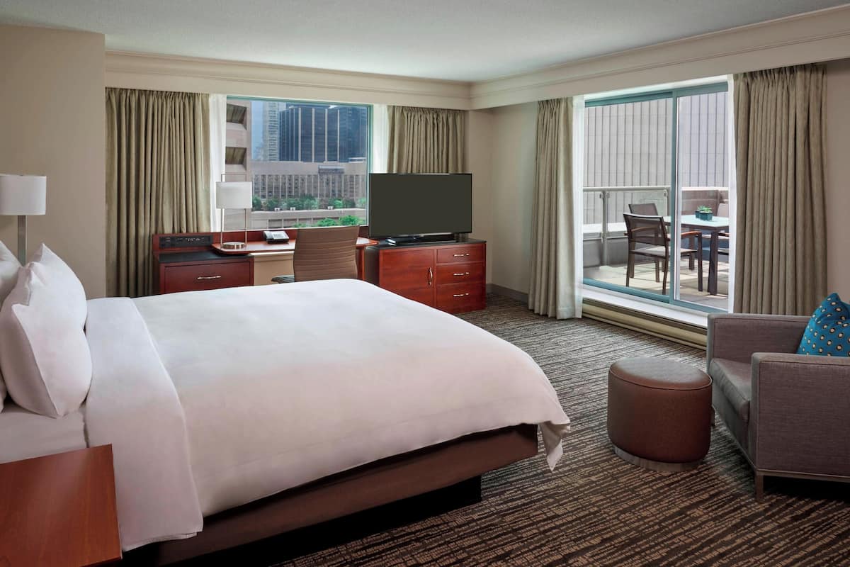 A hotel room with a large bed, a desk with a flat-screen TV, a seating area with a sofa and armchair, and a balcony with a view of the city.